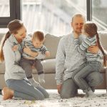 Happy parents with children on knees in casual clothes sitting on floor and playing with kids while spending time together in living room near panoramic window