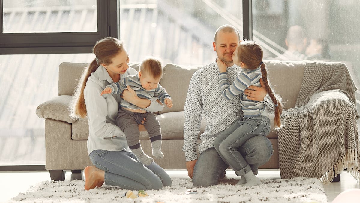 Happy parents with children on knees in casual clothes sitting on floor and playing with kids while spending time together in living room near panoramic window